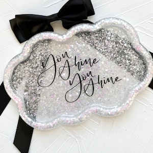 May include: A cloud-shaped tray filled with glitter and the words "You Shine" in black script. The tray has a clear border with a pinkish hue and is accented with a black bow and ribbon.