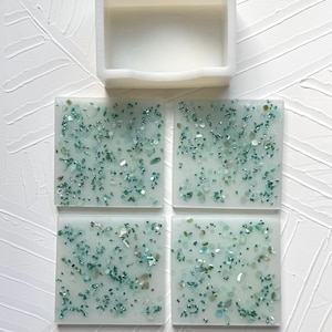 May include: A set of four square coasters with a translucent white base embedded with small, shimmering teal and green glass fragments, accompanied by a matching white square holder for organized storage of the coasters.