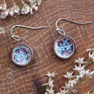 May include: A pair of round earrings with a blue, red, and white speckled design. The earrings have a brown border and silver-coloured hooks. The earrings are displayed on a wooden surface with dried white flowers.