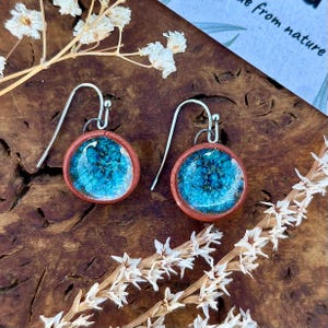 Puede incluir: Pendientes hechos a mano con un diseño redondo turquesa y azul, encerrado en un marco color cobre. Los pendientes tienen ganchos plateados y se exhiben sobre una superficie de madera con flores secas.