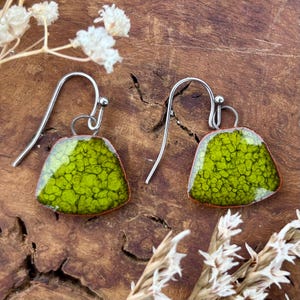 May include: A pair of handmade earrings with a unique design. The earrings feature a silver hook and a green, textured, triangular pendant. The green colour has a speckled pattern. The earrings are displayed on a wooden surface.