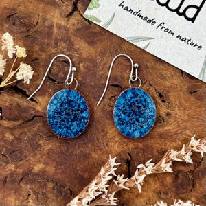 Op de afbeelding: Een paar hangende oorbellen met ronde hangers met een textuur van diepblauw en turquoise kristalpatroon. De oorbellen zijn bevestigd aan zilverkleurige metalen haken. De hangers hebben een gladde, glanzende afwerking en een donkere rand.