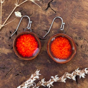 Op de afbeelding: Handgemaakte oorbellen met een gespikkeld oranje en rood ontwerp. De cirkelvormige oorbellen hebben een donkerbruine rand en zilverkleurige oorhaken. De oorbellen worden gepresenteerd op een houten oppervlak. Diameter ongeveer 3 cm.