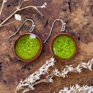 Puede incluir: Un par de pendientes hechos a mano con un centro redondo verde lima y un borde color cobre. Los pendientes tienen un aspecto texturizado y moteado y están suspendidos de ganchos plateados. El fondo es una superficie de madera oscura.