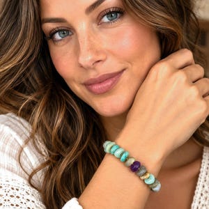May include: A beaded bracelet featuring round, polished stones in shades of turquoise, light gray, and a single deep purple bead, accented with a small, textured gold-toned metal charm. The beads are strung together in a continuous circular design.