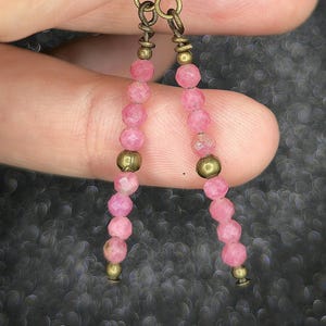 May include: A pair of dangle earrings featuring faceted pink beads strung on a chain with brass-colored accents. The earrings have a gold-colored hook closure. The beads are a light pink color.