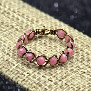 May include: A handmade ring featuring pink gemstone beads woven with gold-colored wire. The ring is displayed on a textured, neutral-toned surface, with a dark, blurred background.