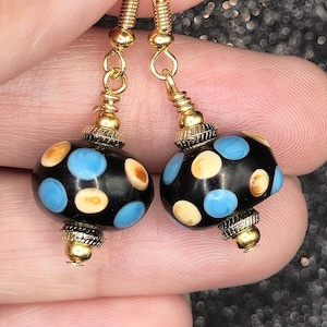 May include: A pair of dangle earrings featuring round black beads with light blue and cream-colored polka dots. The beads are accented with gold-toned metal spacers and caps, suspended from gold-toned metal fishhook ear wires for pierced ears.