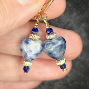 May include: Gold-tone earrings featuring heart-shaped blue and white gemstones. The earrings have a gold hook and are adorned with small blue beads and decorative gold accents. The heart-shaped stones have a marbled appearance.