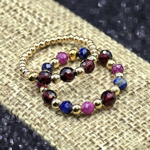 May include: Two beaded rings. One ring is gold with small gold beads. The other ring has gold beads interspersed with dark red, blue, and pink gemstones. The rings are on a tan, textured surface.