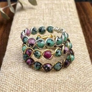 May include: A ring crafted from twisted gold-toned wire featuring a series of small, round, multi-colored beads. The beads display a mix of speckled green, dark grey, and vibrant magenta tones, arranged in a continuous circular band design.