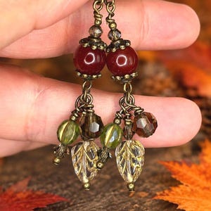 May include: A pair of dangle earrings featuring bronze-toned metal hooks and chains. Each earring displays a large, smooth, deep red spherical bead, accented by smaller faceted olive green and brown beads, and a clear, leaf-shaped glass charm.