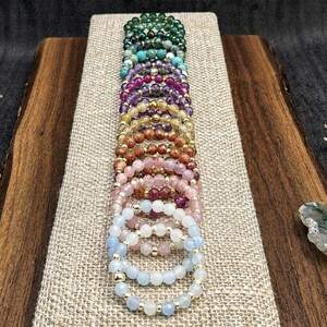 Puede incluir: Una pila vertical de pulseras elásticas de cuentas con varios colores de piedras preciosas, incluyendo verde, turquesa, morado, amarillo, marrón, rosa y azul claro, intercaladas con pequeñas cuentas espaciadoras doradas.