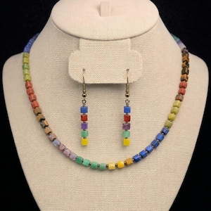 May include: A colorful necklace and earring set displayed on a mannequin. The necklace and earrings feature square beads in a rainbow of colors, including red, orange, yellow, green, blue, and purple. The jewelry is made of small, square beads.