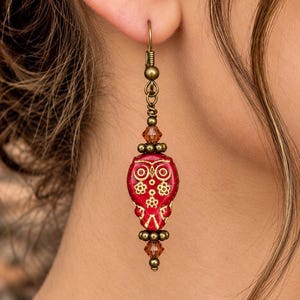 May include: A drop earring featuring a central red glass bead shaped like an owl with gold-colored etched details. The design includes small bronze-toned metal beads and faceted amber-colored crystals above and below the owl, suspended from a hook.
