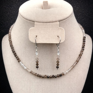 May include: A necklace and earring set displayed on a mannequin. The necklace and earrings feature small, round, brown and clear beads. The necklace is a choker style. The earrings are dangling with a similar bead design.