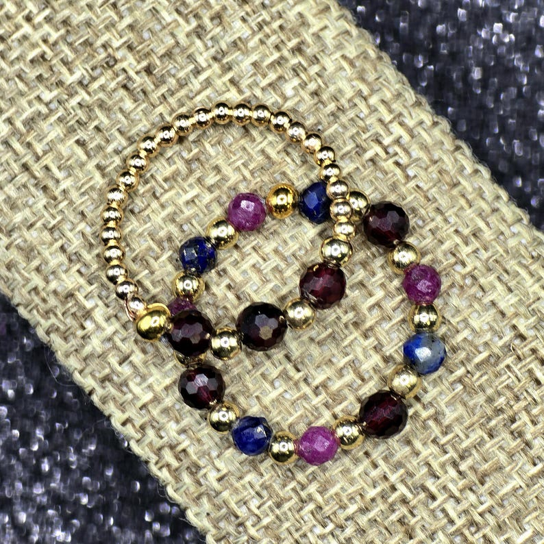 May include: Two gold beaded rings with alternating gemstones. One ring features a pattern of gold beads, purple, blue, and dark red gemstones. The other ring is made of small gold beads. The rings are displayed on a textured, tan surface.