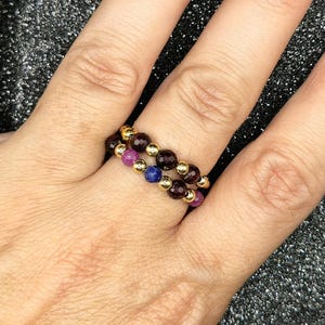 May include: Two beaded rings on a finger. One ring features alternating gold beads and dark red beads. The other ring has gold beads alternating with purple and blue beads. The rings are worn on a finger.