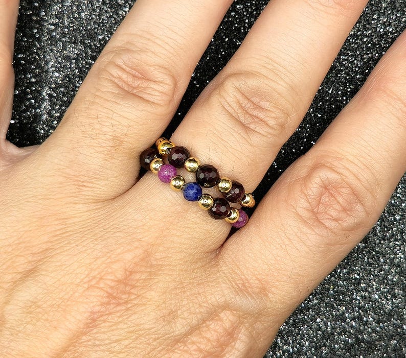 May include: Two beaded rings on a finger. One ring features alternating gold beads and dark red beads. The other ring has gold beads alternating with purple and blue beads. The rings are worn on a finger.