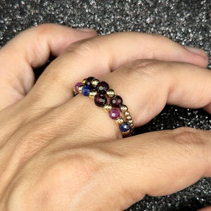May include: Two beaded rings with gold-colored metal and colorful gemstones. One ring features dark red, purple, and gold beads. The other ring has dark red, blue, and gold beads. The rings are on a hand.