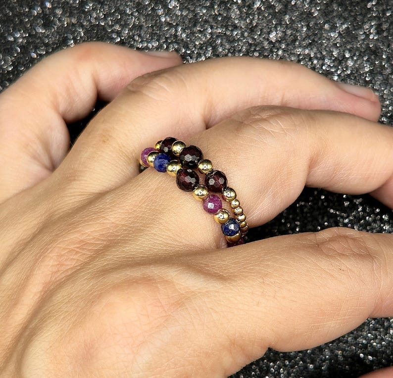 May include: Two beaded rings with gold-colored metal and colorful gemstones. One ring features dark red, purple, and gold beads. The other ring has dark red, blue, and gold beads. The rings are on a hand.