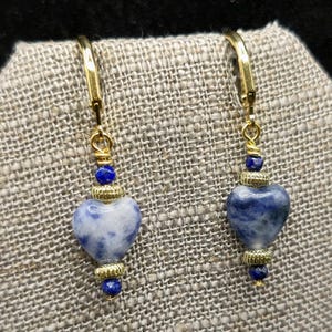 May include: A pair of gold-colored leverback earrings. Each earring features a heart-shaped blue and white stone, accented with gold-colored beads and small blue beads. The earrings are displayed on a textured, neutral-colored surface.