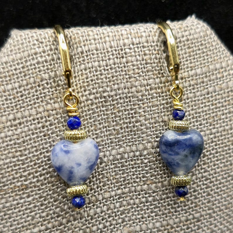 May include: A pair of gold-colored leverback earrings. Each earring features a heart-shaped blue and white stone, accented with gold-colored beads and small blue beads. The earrings are displayed on a textured, neutral-colored surface.