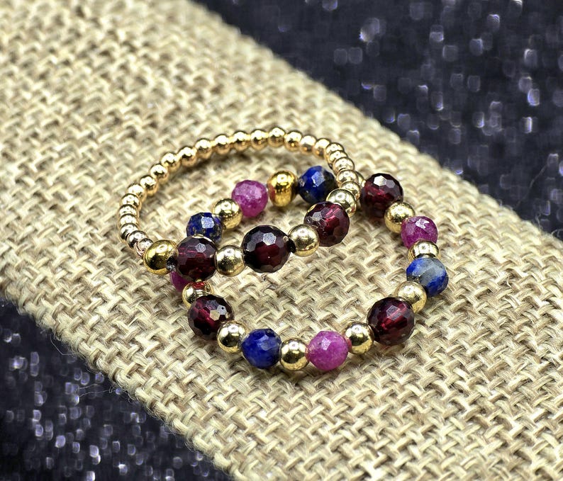 May include: Two beaded rings. One ring is gold with small gold beads. The other ring has gold beads interspersed with dark red, blue, and pink gemstones. The rings are on a tan, textured surface.