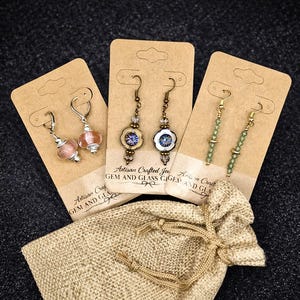 May include: Three pairs of artisan earrings on display cards, alongside a small burlap bag. The earrings feature various designs, including beaded and glass elements. The cards have the text "Artisan Crafted Gem and Glass".