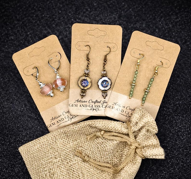 May include: Three pairs of artisan earrings on display cards, alongside a small burlap bag. The earrings feature various designs, including beaded and glass elements. The cards have the text "Artisan Crafted Gem and Glass".