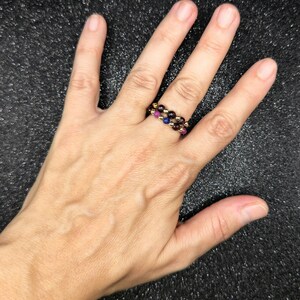 May include: Two beaded rings with a gold band. One ring features alternating red and gold beads, while the other has red, blue, and gold beads. The rings are worn on a hand against a dark background.