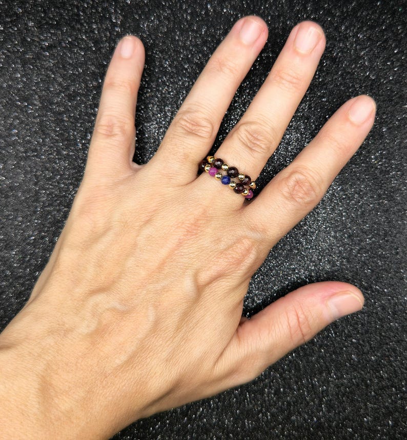 May include: Two beaded rings with a gold band. One ring features alternating red and gold beads, while the other has red, blue, and gold beads. The rings are worn on a hand against a dark background.
