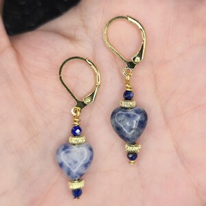 May include: A pair of gold-colored leverback earrings. Each earring features a heart-shaped, blue and white stone pendant, accented with gold-colored beads and a small blue bead. The earrings are designed for pierced ears.