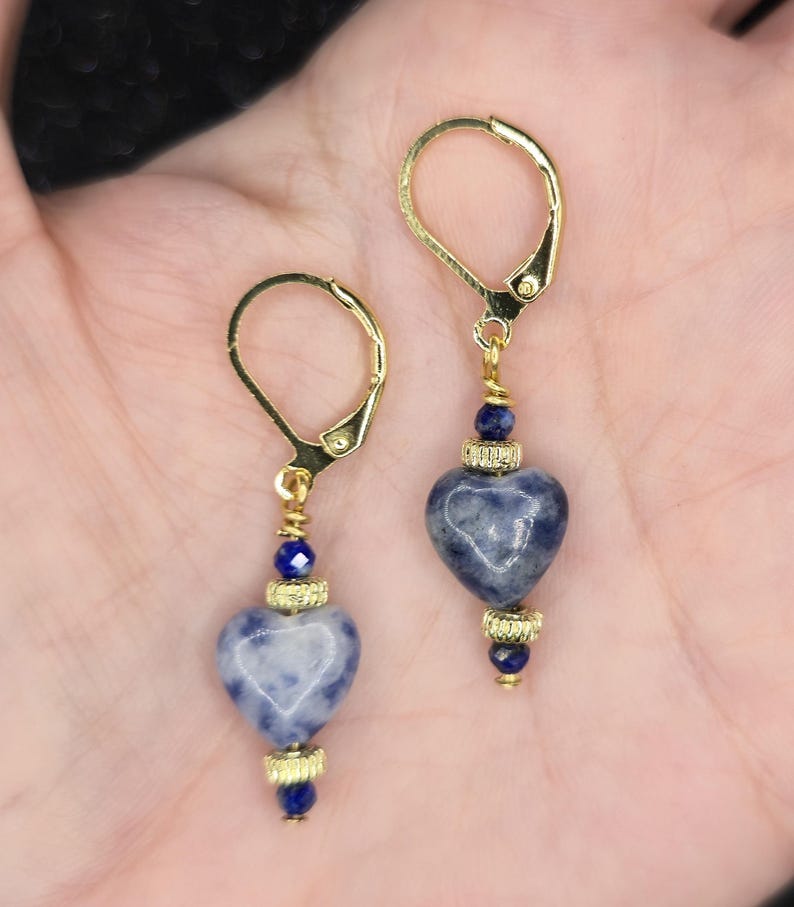 May include: A pair of gold-colored leverback earrings. Each earring features a heart-shaped, blue and white stone pendant, accented with gold-colored beads and a small blue bead. The earrings are designed for pierced ears.