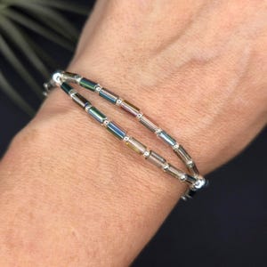 May include: A double-strand bracelet featuring iridescent, rectangular beads. The beads display a range of colors, including green, blue, and gold, and are strung on a silver chain. The bracelet is designed to wrap around the wrist.