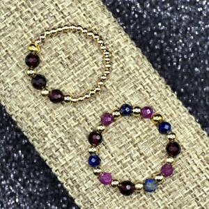 May include: Two beaded rings. One ring is gold with dark red beads. The other ring is gold with a mix of dark red, purple, and blue beads. The rings are displayed on a tan, textured surface.
