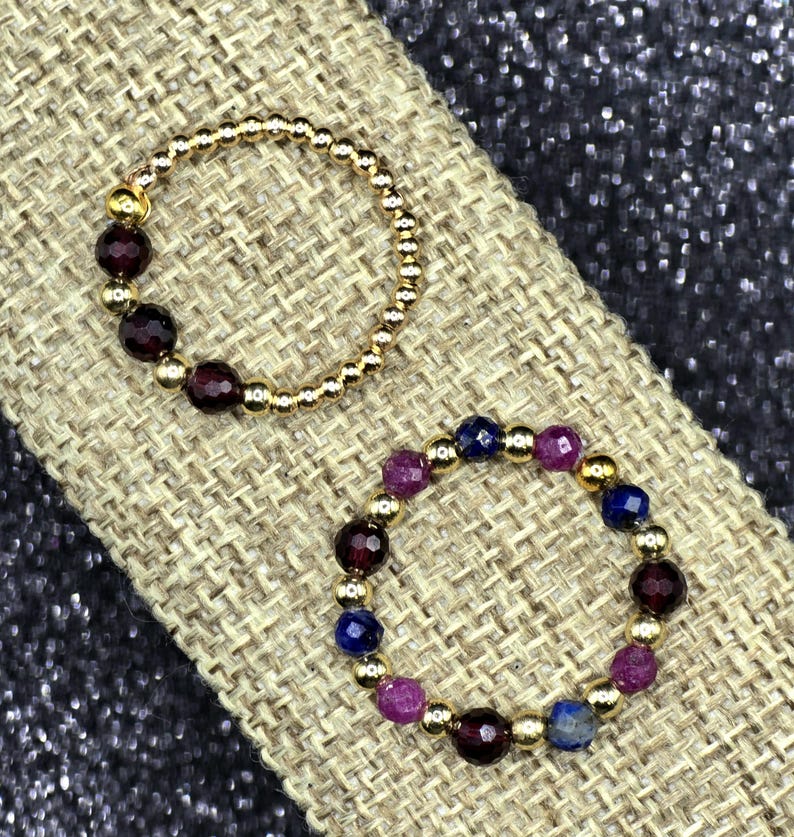 May include: Two beaded rings. One ring is gold with dark red beads. The other ring is gold with a mix of dark red, purple, and blue beads. The rings are displayed on a tan, textured surface.