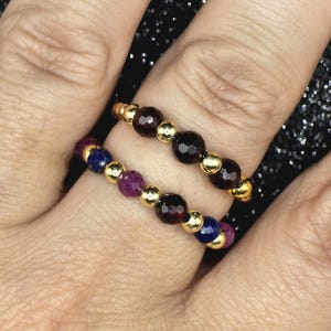 May include: Two beaded rings with gold-colored accents. One ring features dark red beads, while the other has a combination of purple, blue, and red beads. The rings are worn on a finger.