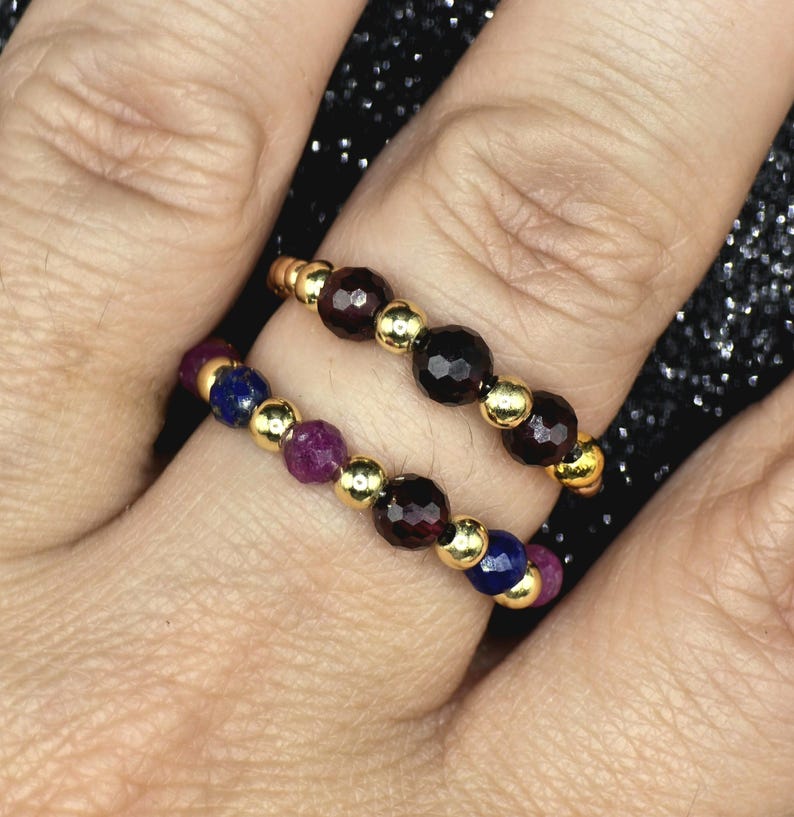 May include: Two beaded rings with gold-colored accents. One ring features dark red beads, while the other has a combination of purple, blue, and red beads. The rings are worn on a finger.