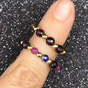 May include: Two beaded rings with gold accents. One ring features dark red beads, while the other combines dark red, pink, and blue beads. The rings are displayed on a finger against a sparkly, dark background.