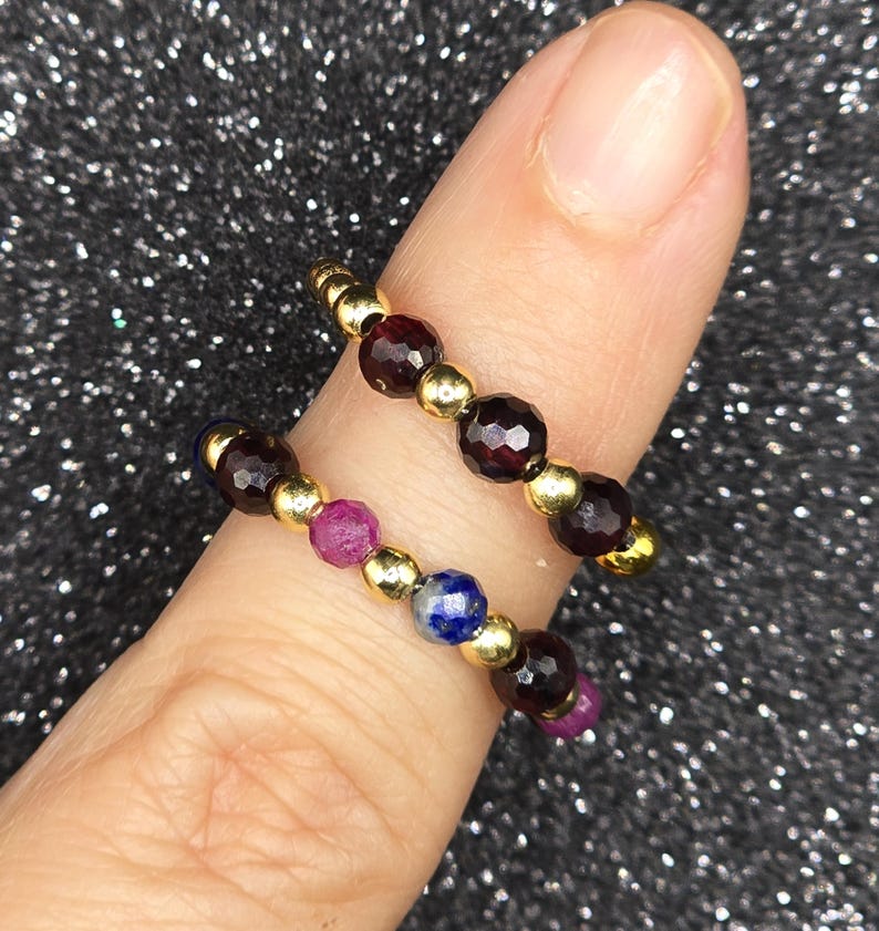 May include: Two beaded rings with gold accents. One ring features dark red beads, while the other combines dark red, pink, and blue beads. The rings are displayed on a finger against a sparkly, dark background.