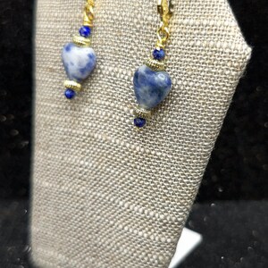 May include: A pair of gold-colored earrings with heart-shaped blue and white stones. The earrings have a lever back closure and are displayed on a textured, light-colored surface. Small blue beads accent the design.