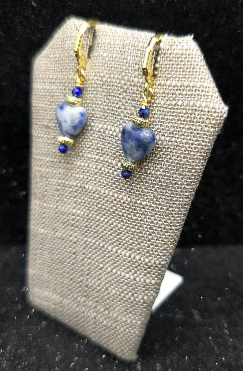 May include: A pair of gold-colored earrings with heart-shaped blue and white stones. The earrings have a lever back closure and are displayed on a textured, light-colored surface. Small blue beads accent the design.