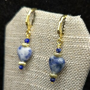 May include: A pair of gold-colored leverback earrings. Each earring features a heart-shaped blue and white stone, accented with gold-colored beads and a small blue bead at the bottom. The earrings are displayed on a textured gray surface.