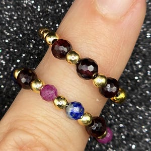 May include: Two beaded rings with gold-colored beads and faceted gemstones. One ring features deep red garnet stones, while the other has a combination of red, blue, and purple gemstones. The rings are displayed on a finger.