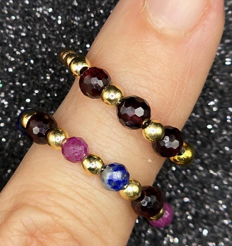 May include: Two beaded rings with gold-colored beads and faceted gemstones. One ring features deep red garnet stones, while the other has a combination of red, blue, and purple gemstones. The rings are displayed on a finger.