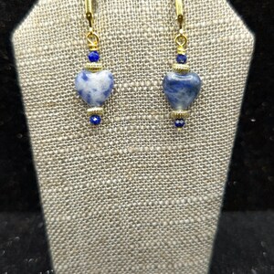 May include: A pair of gold-toned earrings with heart-shaped blue and white stone pendants. The earrings have gold-colored hooks and are accented with small blue beads. The earrings are displayed on a textured, neutral-colored surface.