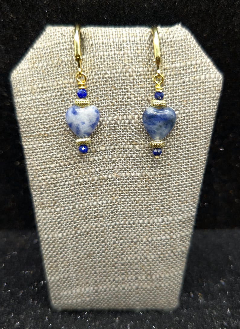 May include: A pair of gold-toned earrings with heart-shaped blue and white stone pendants. The earrings have gold-colored hooks and are accented with small blue beads. The earrings are displayed on a textured, neutral-colored surface.
