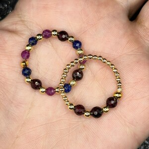 May include: Two beaded rings with gold-colored beads and gemstones. One ring features a combination of dark red, purple, and blue gemstones. The other ring has dark red gemstones. The rings are displayed on a light-colored surface.