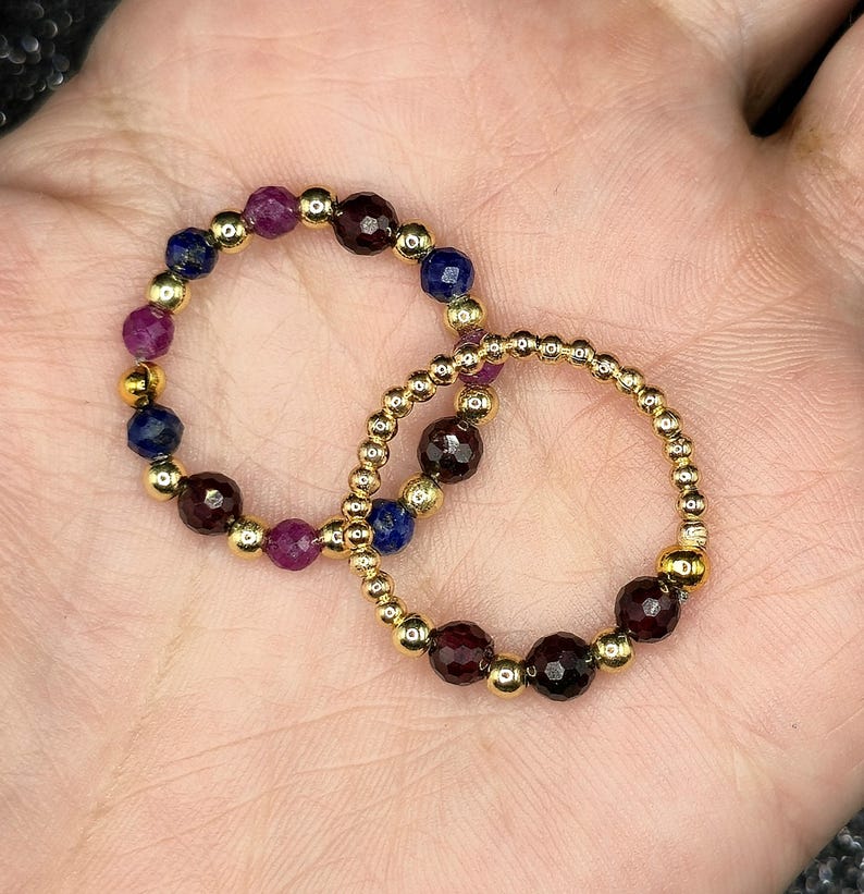 May include: Two beaded rings with gold-colored beads and gemstones. One ring features a combination of dark red, purple, and blue gemstones. The other ring has dark red gemstones. The rings are displayed on a light-colored surface.